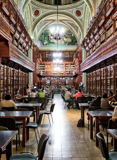Grand library reading room with tall wooden shelves, arched ceiling, chandeliers, and people seated at long tables.