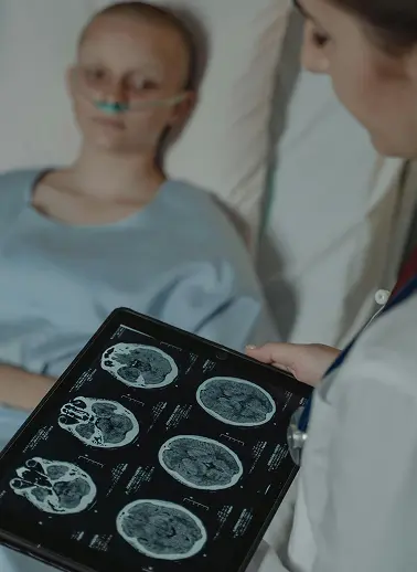 Doctor reviews brain MRI scans on a tablet beside a patient in a hospital bed wearing oxygen mask.