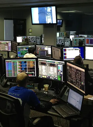 Mission control room with teams at desks surrounded by many computer monitors displaying flight data and charts.